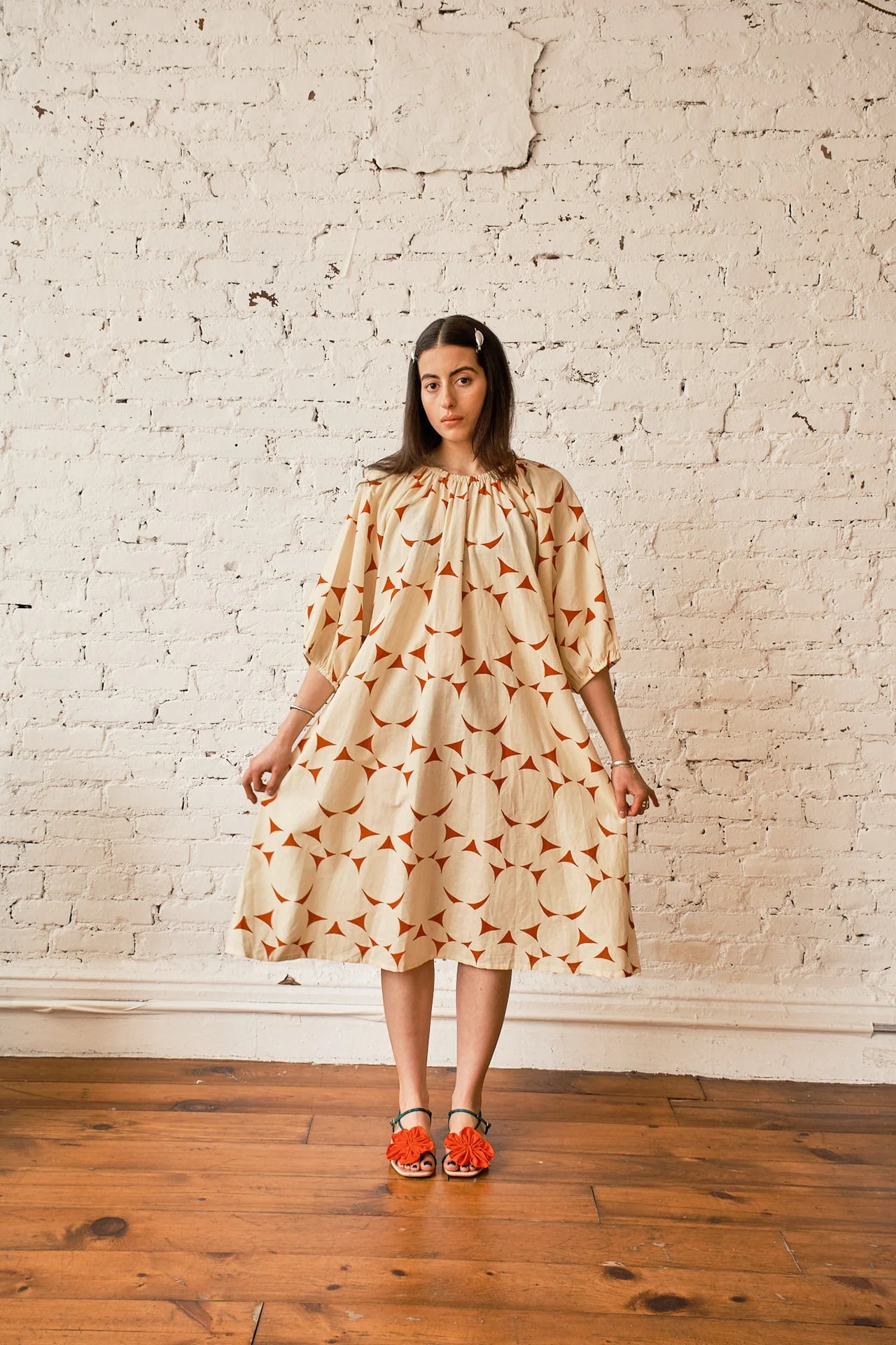 A woman modeling a cream-colored midi dress with an orange geometric pattern, standing on a wooden floor against a white brick wall.