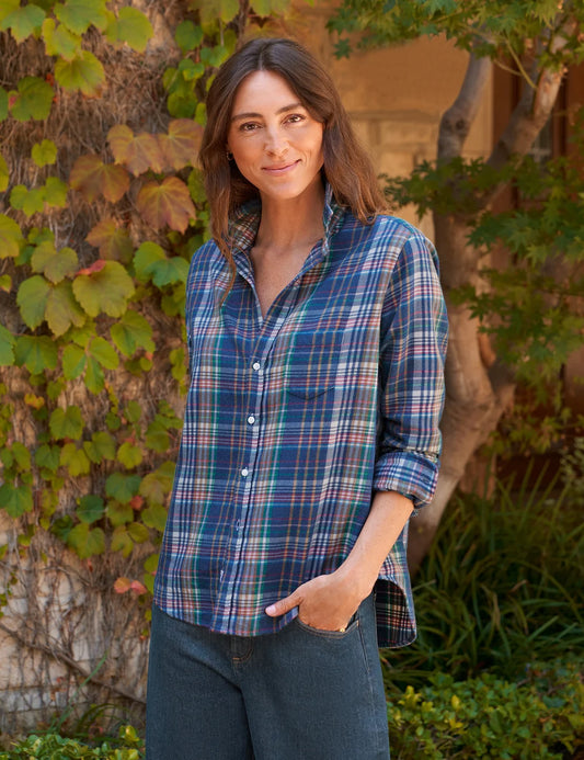 A woman wearing a blue and green plaid button-down shirt with dark jeans, standing outdoors in front of a leafy vine-covered wall.