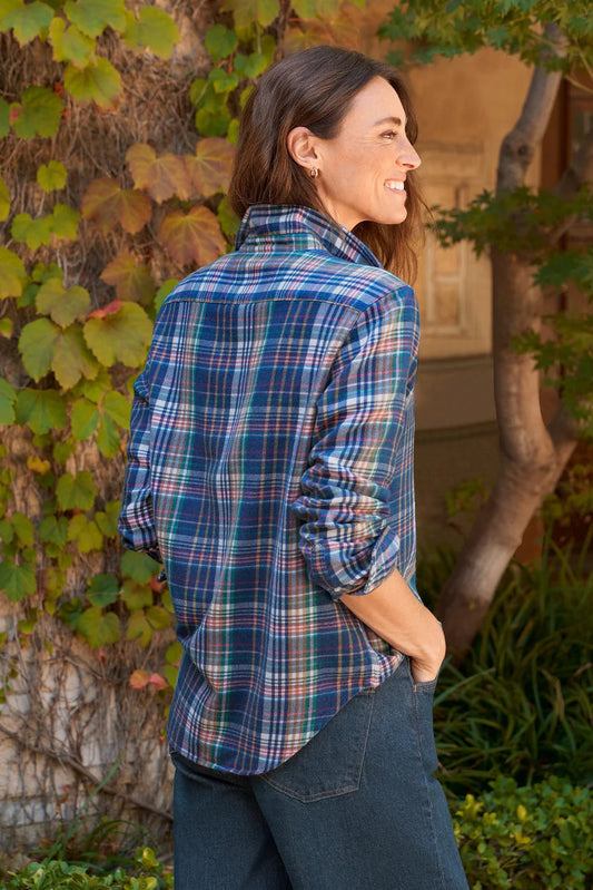 A woman wearing a blue and green plaid button-down shirt with dark jeans, standing outdoors in front of a vine-covered wall.