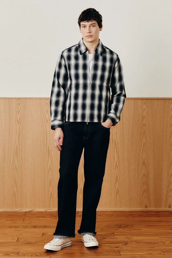 A man stands in a studio setting wearing a black and white plaid zip-up overshirt, dark navy pants, and white sneakers, with a wood-paneled wall and wooden floor behind him. - 2
