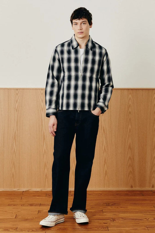 A man stands in a studio setting wearing a black and white plaid zip-up overshirt, dark navy pants, and white sneakers, with a wood-paneled wall and wooden floor behind him.