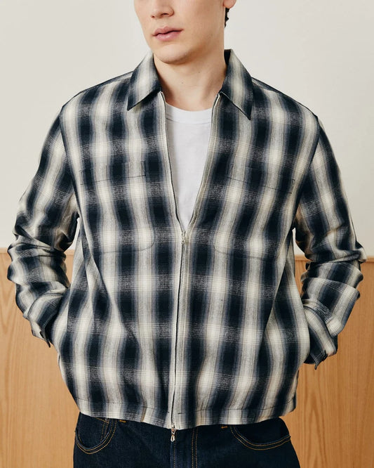 A man wearing a dark navy and off-white plaid zip-up overshirt over a white t-shirt and dark jeans, shown from the chin down against a neutral indoor background.