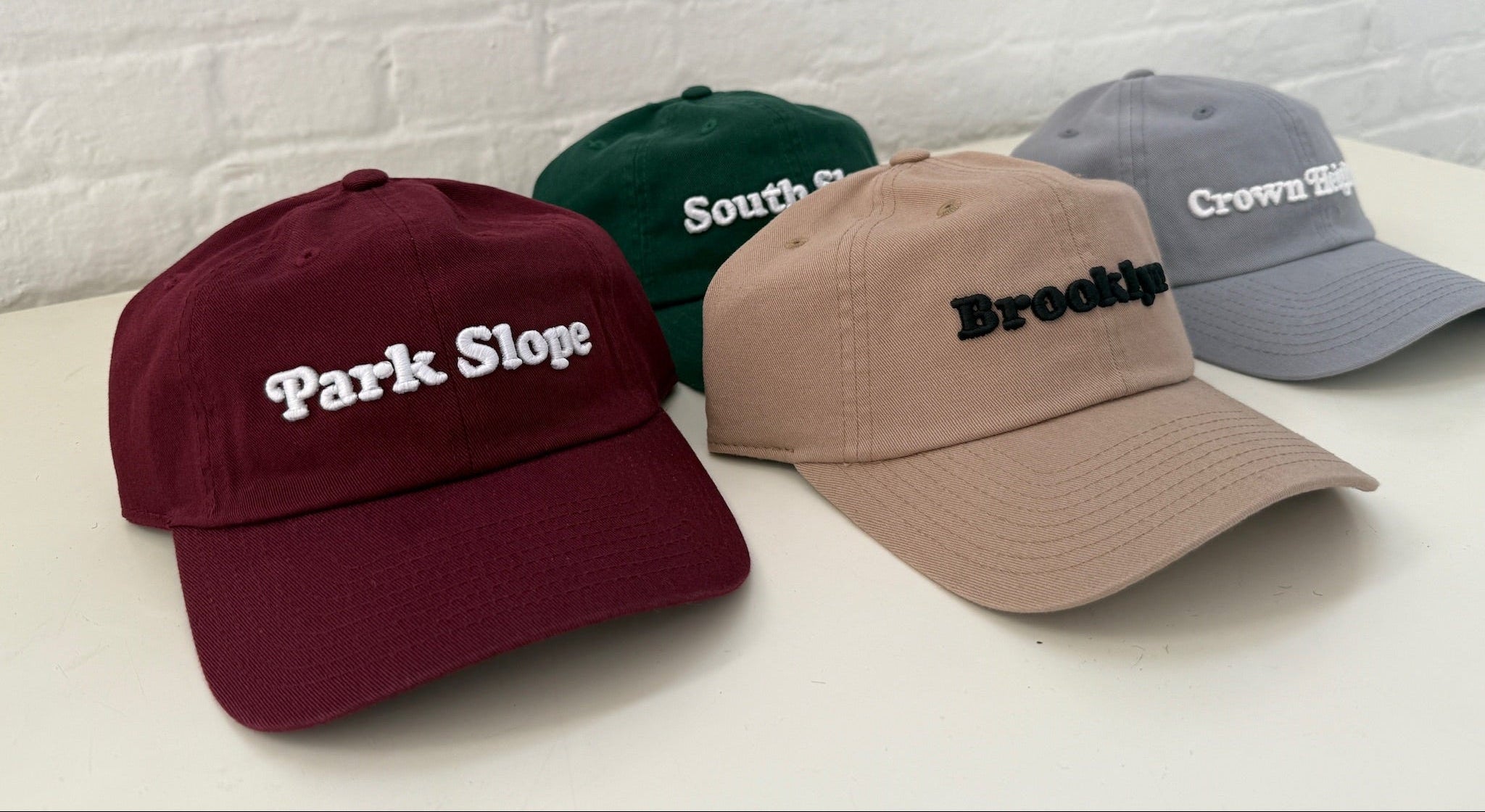 Four baseball caps in maroon, green, beige, and gray, arranged on a white surface against a white brick wall. Each cap has embroidered text on the front: 'Park Slope' in white, 'South Slope' in white, 'Brooklyn' in black, and 'Crown Hill' in white.