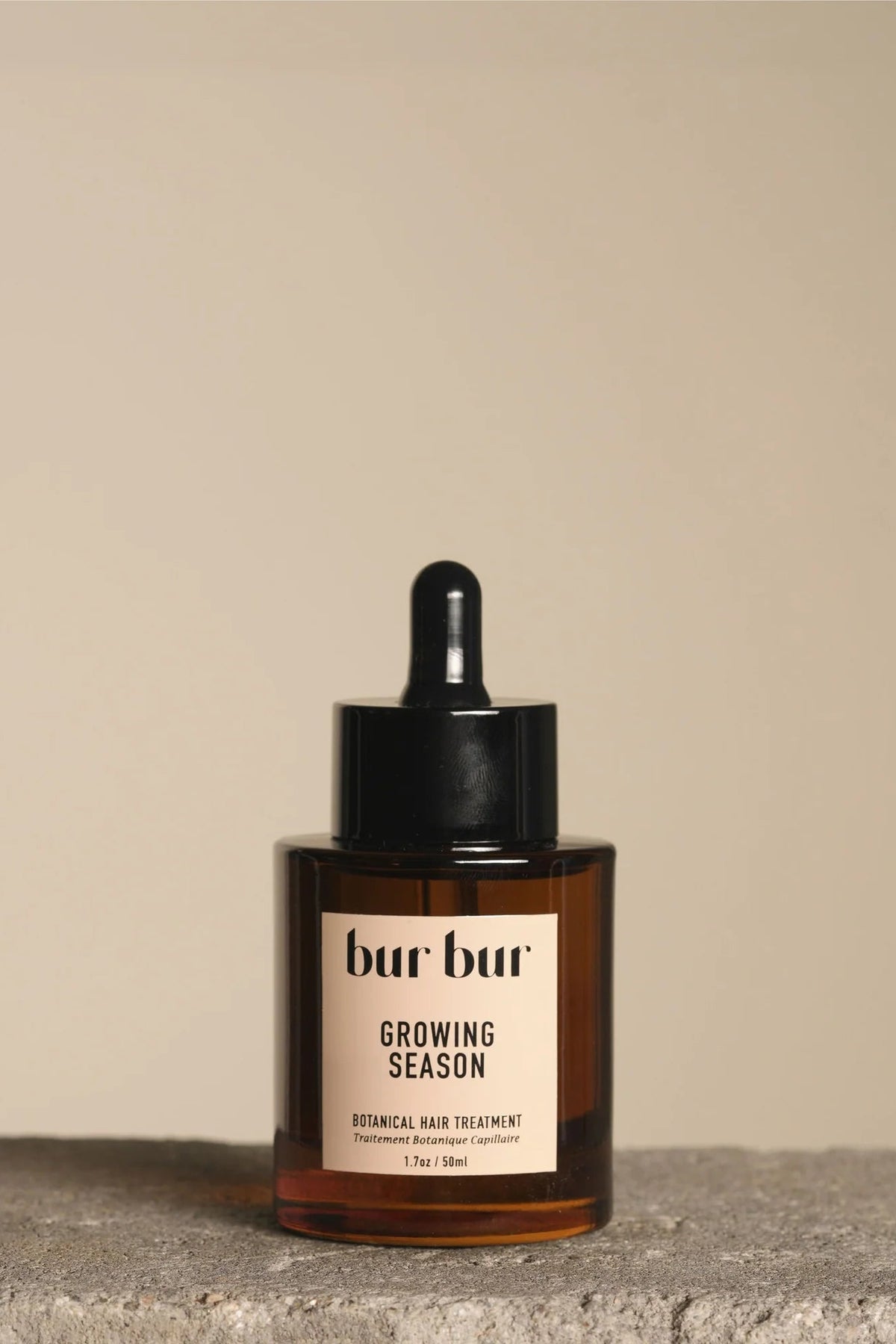 A bottle of bur bur Growing Season Botanical Hair Treatment with a black dropper cap and cream-colored label, set on a textured stone surface against a solid beige background.