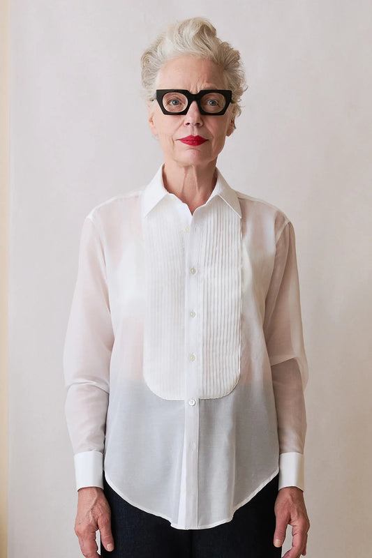 An older woman with short gray hair and black-rimmed glasses wears a white long-sleeve button-up blouse with a pleated front panel over a sheer white underlayer, paired with dark pants, against a plain light beige wall.