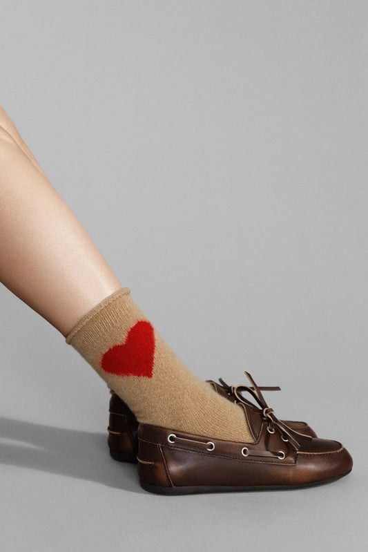 Beige sock with a red heart design worn by a person with brown shoes on a gray background